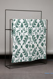 White blanket with green floral pattern draped over a black metal frame against a gray wall.
