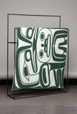 Decorative blanket with abstract green and white pattern on a black metal stand against a gray wall.
