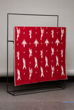 Red blanket with white patterns on a black stand against a gray wall.