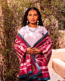 A woman stands outdoors in front of a warm adobe wall and greenery, wearing a light pink top and a draped magenta and teal patterned shawl or wrap. She is also wearing turquoise jewelry.