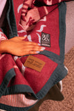 A close-up of a red and white patterned blanket with dark gray trim, featuring a black '8th GEN' brand patch and a tan leather patch, with a hand resting on it.