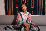 A woman sits on a gray couch wearing a white t-shirt and a pink, teal, and red patterned shawl. She is wearing turquoise beaded necklaces and earrings.