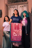 Three women standing in front of a rustic wooden door, holding up a pink and teal patterned textile blanket. The center woman wears a navy blue t-shirt.