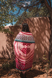 A person standing outdoors in front of a tan adobe wall under tree shadows, wrapped in a patterned blanket. The blanket has a pink and white Southwestern-style design with a teal border.