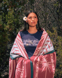 A woman outdoors wearing a navy blue graphic t-shirt and a pink and white patterned blanket draped over her shoulders, with green foliage in the background.