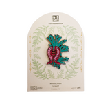 An enamel pin shaped like a bleeding heart, displayed on its original arched backing card. The pin features a pink heart-shaped flower with green leafy tops and red accents, outlined in black.