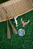 A set of outdoor-themed souvenirs displayed on artificial green grass next to a wooden canoe and paddles. The pins include a black and white fish with a red and white striped hook, a red deer head with antlers, and a circular magnet featuring the 'Snoqualmie Falls' landscape design.