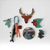 A set of colorful enamel pins and a keychain with a carabiner, arranged on a plain white background. The pins feature local wildlife and landscape designs, including an elk head, a fish, a waterfall, two berries, and a flower.