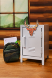 A miniature white metal safe or locker with an elk head emblem on the door, displayed on a wooden surface next to a small camouflage pouch. The background shows a warm wood-paneled wall and a window.