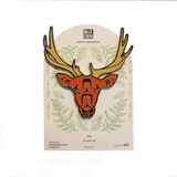 An enamel pin shaped like a stylized elk head with antlers, mounted on its backing card. The pin features a rust-orange face with black outlines and gold antlers.