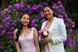 Two women standing in front of purple flowers, one in a pink dress and the other in a white suit.