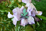 A close-up of an enamel pin resting on the center of a purple flower, set against a background of green foliage.