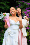 Two women, one in a white dress holding flowers and the other in a light-colored outfit, standing outdoors with greenery and purple flowers in the background.