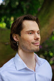 Man with a beard wearing a light blue shirt outdoors with greenery in the background