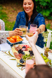 People toasting with glasses at a outdoor gathering with food and drinks.