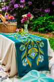 Tablecloth with floral design on a table outdoors with a picnic basket and flowers.