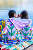 Two people sitting by a lake, wrapped in a colorful towel with leaf patterns.