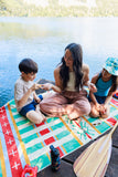 Woman and two children sitting on a colorful mat by a lake with a paddle and bottle nearby.