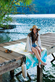 Woman sitting on a dock by a lake, wearing a hat and light clothing.