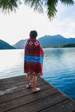 Person wrapped in a red blanket with blue patterns standing on a wooden dock by a lake with mountains in the background.