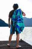 Person standing on a dock with a blue and green towel draped over their shoulder, looking out at water and mountains.