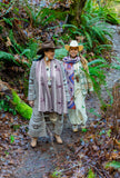 Two women walking side by side on a mossy forest path surrounded by green ferns wearing geometric patterned wool scarves