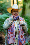 A woman wearing a geometric patterned knit wool scarf with purple, rust red, and cream maple leafs in a green forest