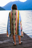 Person wearing a colorful patterned towel standing on a wooden dock by a lake with mountains in the background