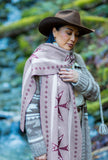 A person wearing a gray knit sweater with a dusty grey geometric patterned knot wool scarf with dark purple geometric stars, standing outdoors in a green forest