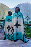 Two people standing on a wooden deck with their backs to the camera, wrapped in matching turquoise, cream, and black patterned blankets, overlooking a scenic lake and mountains.