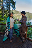 Two men standing on a wooden deck outdoors by a lake, wrapped in and holding a woven blanket with turquoise, black, cream, and brown stripes. Trees and a mountain are visible in the background.