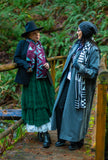 Two people standing on a wooden bridge in a forest, dressed in long coats and hats, modeling geometric patterned knit wool scarves