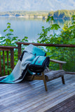 A teal, black, and white fleece blanket and matching backpack are draped over a wooden chair on a wooden deck, with a scenic lake and forest in the background.