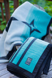 A folded and draped blanket with a teal, white, and black star design, resting on a wooden deck. A small black brand label is visible on the front.
