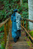 A person viewed from behind walking on a rustic wooden boardwalk bridge in the forest with a black and white geometric patterned wool scarf 
