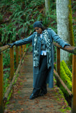 A person posing on a rustic wooden bridge in a mossy forest, wearing a long slate blue coat and a black and white geometric patterned wool scarf hanging