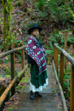 A woman stands on a wooden bridge in a forest, wearing a geometric patterned knit wool scarf as a shawl with geometric flowers in dark lilac, cream, and bright light blue.