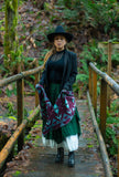 A woman stands on a wooden bridge in a forest, wearing a geometric patterned knit wool scarf as a wrap with geometric flowers in dark lilac, cream, and bright light blue.