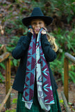 A woman wearing a black wide-brim hat, black coat, and green skirt stands on a wooden bridge in a forest, wearing a geometric patterned knit wool scarf with geometric flowers in dark lilac, cream, and bright light blue.