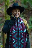 A woman outdoors wearing a wide-brim black hat wearing a geometric patterned knit wool scarf with geometric flowers in dark lilac, cream, and bright light blue.