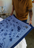 A person holds up a light blue blanket with a dark blue geometric pattern and a dark blue border. The blanket has a small black woven label.