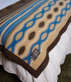 A close-up of a patterned blanket draped over white bedding. The blanket features a beige base with wavy blue and brown geometric patterns, a dark brown border, and a visible brand label.
