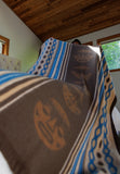A close-up of a patterned blanket or throw draped in a bedroom, showing a brown base with tan face motifs and blue, beige, and black geometric border stripes. A person is partially visible behind the fabric near a window.