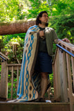 A man wearing glasses stands on wooden outdoor stairs wrapped in a patterned blanket. The blanket features a cream base with blue, brown, and beige geometric wave-like patterns.