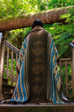 A person viewed from behind wearing a large woven blanket with a brown center panel featuring repeating moon face motifs and blue-and-beige striped sides, standing on wooden steps in a forest setting.