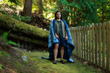 A man standing on a mossy forest path wearing a blue patterned blanket draped over his shoulders, with a wooden fence and trees in the background.