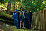 A man standing outdoors in a wooded area next to a wooden fence, holding up a dark blue patterned blanket or scarf spread wide like a cape. He is wearing a green shirt, gray t-shirt, and dark shorts.