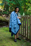 A man standing outdoors in a wooded area next to a wooden fence, wrapped in a light blue blanket with a dark blue geometric pattern.