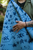 A close-up of a person wrapped in a light blue blanket with a dark blue geometric pattern, wearing an olive green shirt outdoors.