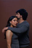 A man and a woman in a studio setting, with the man wearing a dark gray long-sleeve knit sweater and the woman wearing a matching dark gray sleeveless top. The man has curly dark hair and a beard, and the woman has long dark hair.
